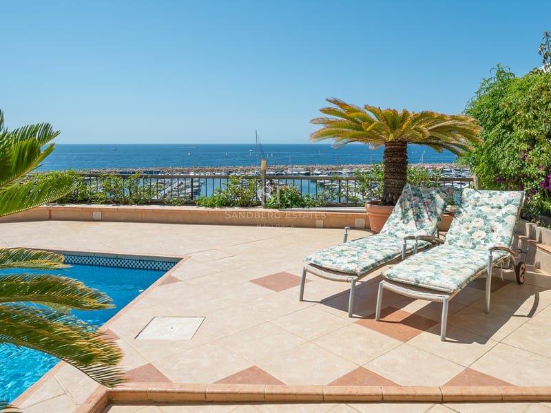 Terrasse surélevée avec piscine, chaises longues et vue panoramique sur un port de plaisance et la mer Méditerranée.