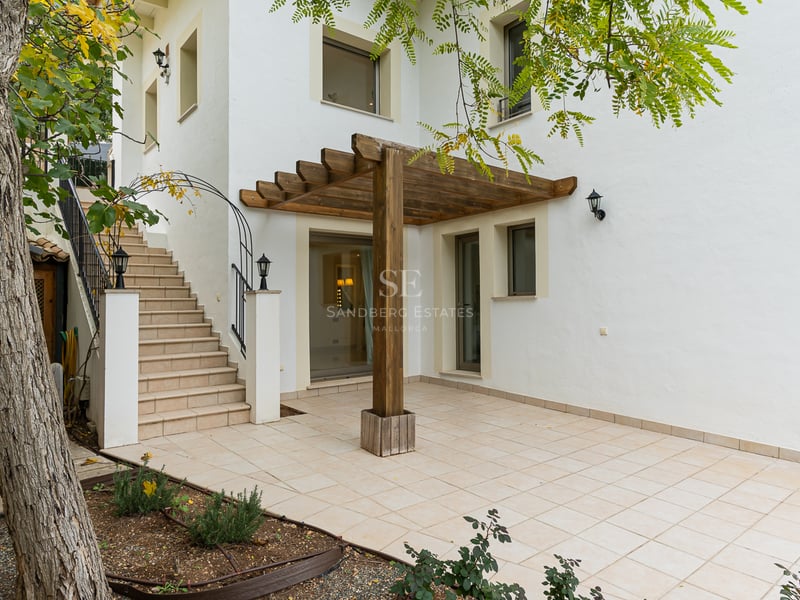 Extérieur de bâtiment blanc avec patio carrelé, pergola en bois et escaliers en pierre menant à l'étage.