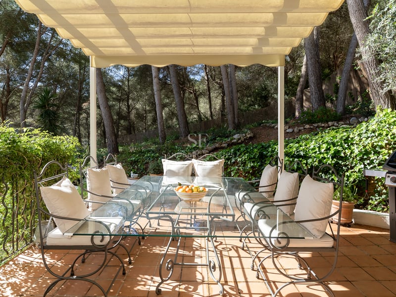 Terrasse avec table en fer forgé, plateau en verre, coussins crème, entourée d'une pinède luxuriante.