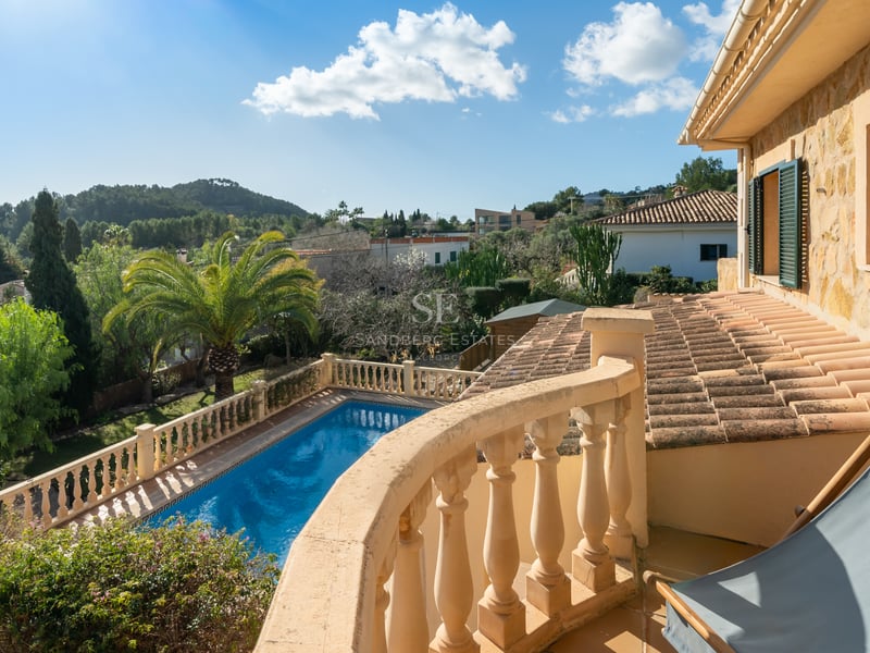 Vue d'un balcon en pierre sur une piscine bleue, des jardins luxuriants et des collines sous un ciel dégagé.