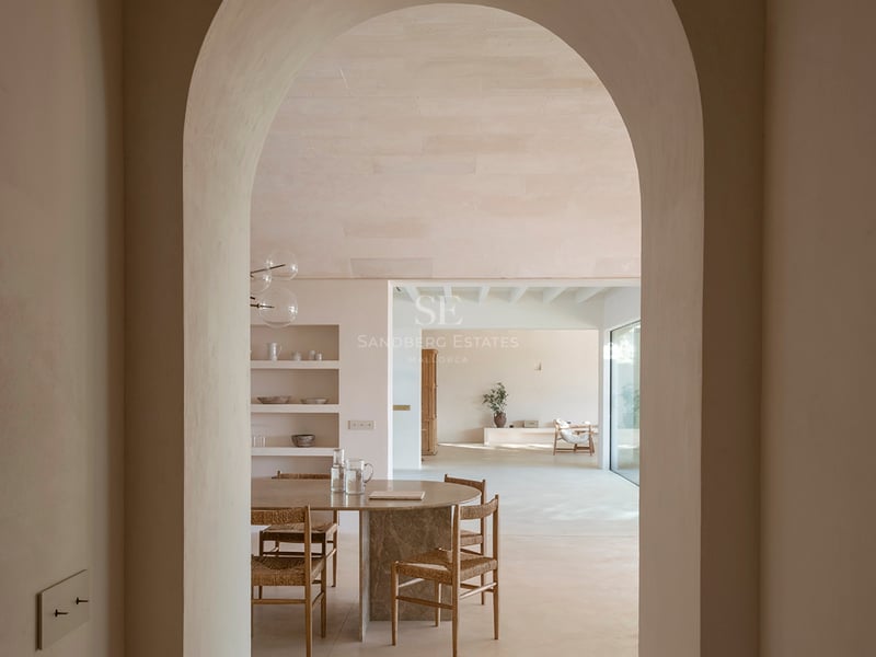 View through a plaster archway into a dining area featuring a stone table, wicker chairs, and sand-colored microcement.