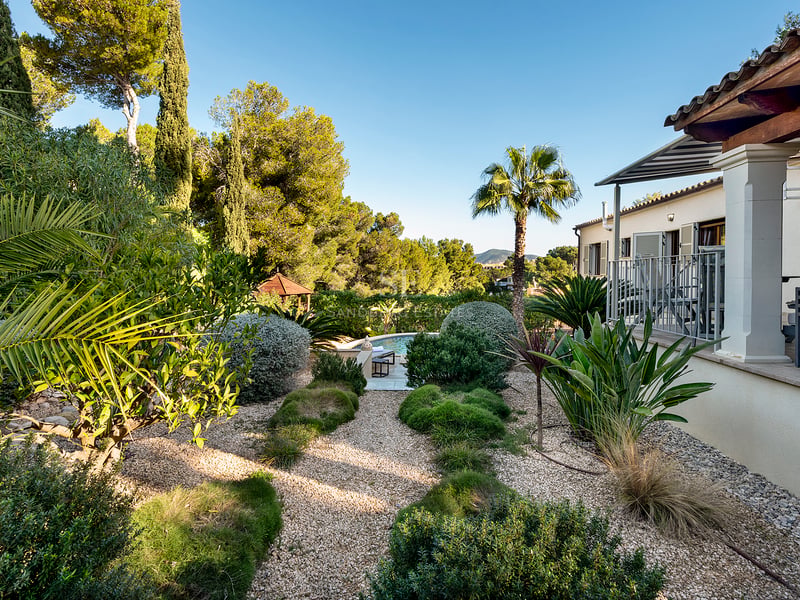 Jardin paysager luxuriant avec allées de gravier menant à une piscine, à côté d'une villa de style méditerranéen.