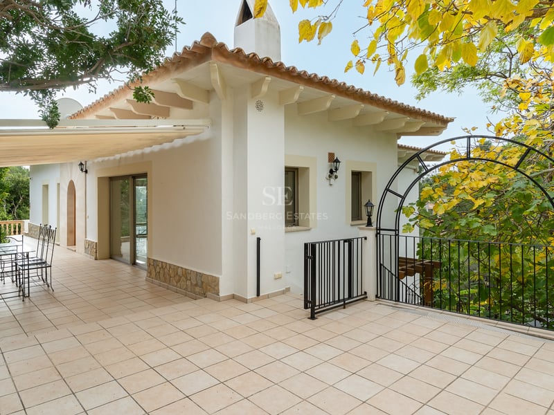 Grande terrasse carrelée avec mobilier en fer forgé, façade blanche et végétation luxuriante sous un auvent.