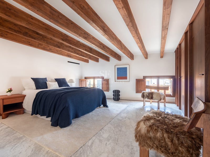 Dormitorio de lujo con vigas de madera oscura, suelo de mármol blanco, ropa de cama azul marino y taburetes de piel.