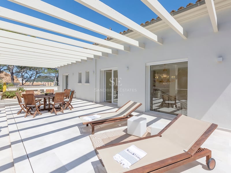 Terrasse blanche lumineuse avec pergola en bois, table à manger extérieure et chaises longues sous un ciel bleu.