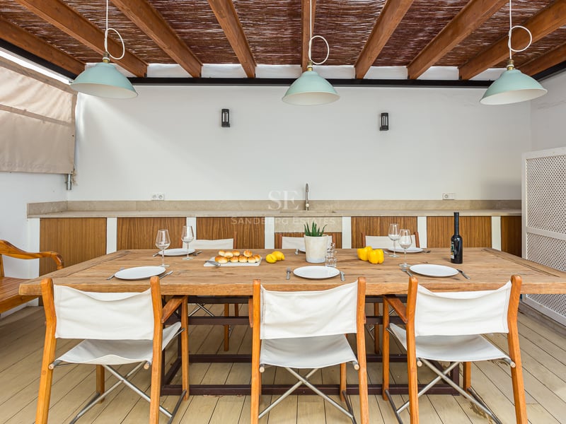 Terrasse couverte avec grande table en bois, chaises en toile blanche et cuisine extérieure sous une pergola.