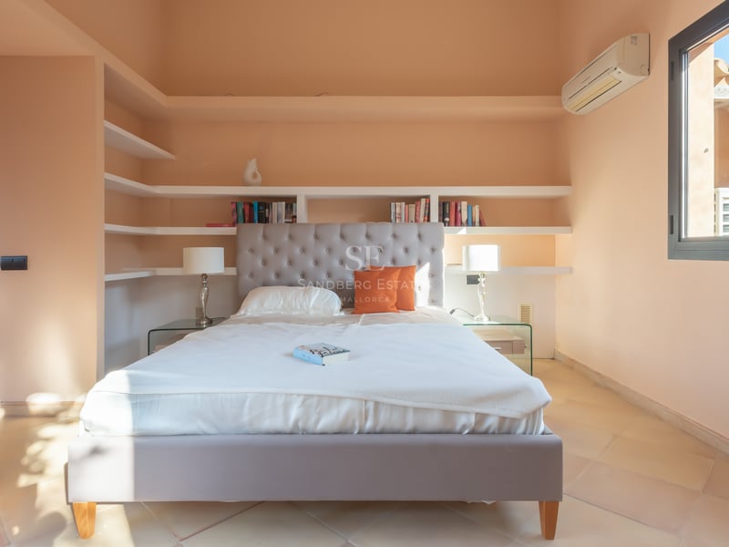 Chambre lumineuse avec tête de lit capitonnée grise, literie blanche, étagères encastrées et sol en terre cuite.
