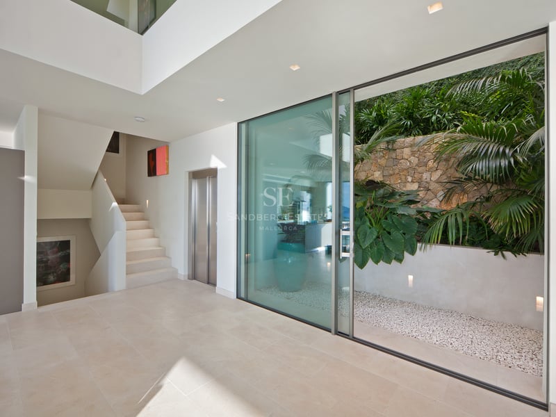Hall minimaliste avec sol en pierre claire, ascenseur, escalier blanc et baie vitrée donnant sur un patio tropical.