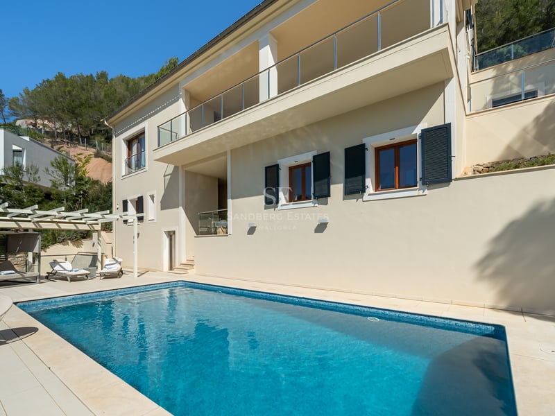 Piscine de luxe à côté d'une villa de couleur crème avec balcons en verre et forêt de pins en arrière-plan.