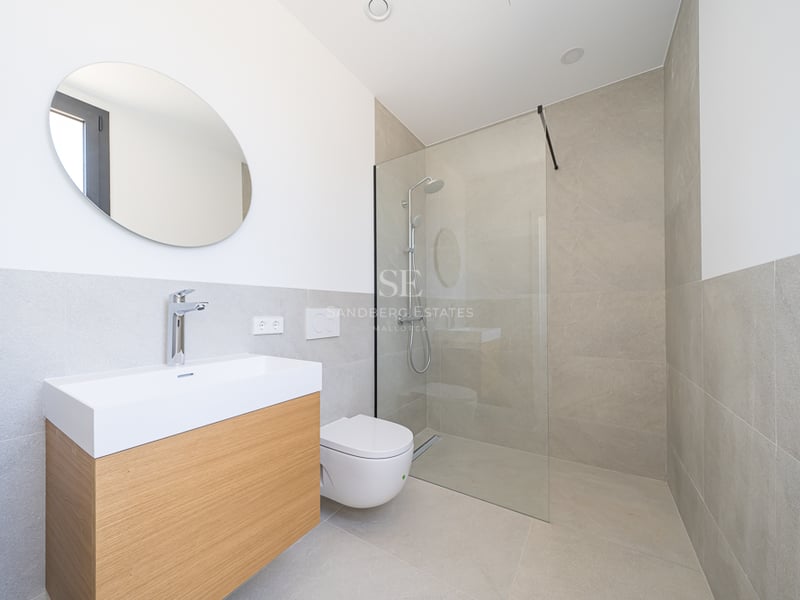 Salle de bain minimaliste avec meuble en bois clair, miroir rond, WC blanc et douche en verre avec carrelage beige.