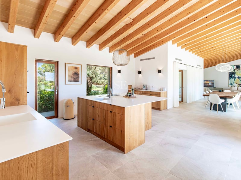 Cuisine spacieuse avec îlot en chêne, plans de travail blancs, poutres apparentes au plafond et sol en pierre.