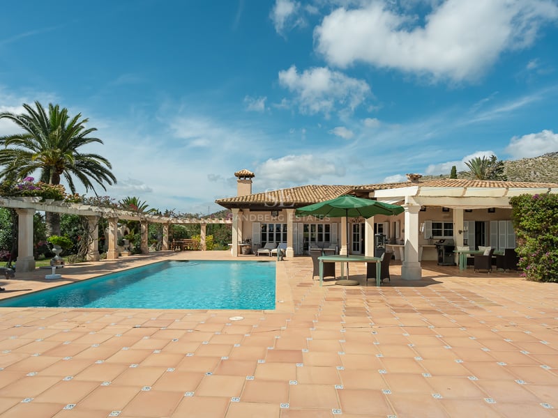 Large rectangular swimming pool surrounded by a terracotta terrace and a white villa under a clear blue sky.