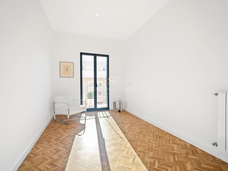 Pièce blanche lumineuse avec parquet en bois en chevrons, fauteuil blanc et porte-fenêtre donnant sur un balcon.
