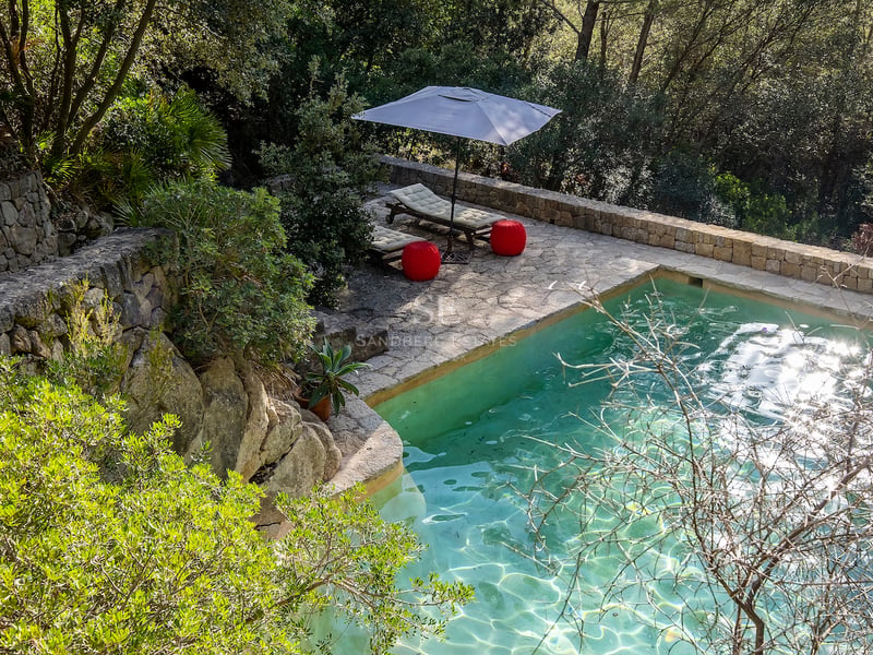 Vue surélevée d'une piscine turquoise avec terrasse en pierre, chaises longues et poufs rouges dans une forêt.