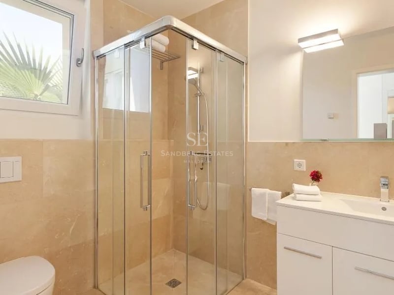 Salle de bain lumineuse avec douche d'angle en verre, carrelage en marbre beige et meuble vasque blanc.