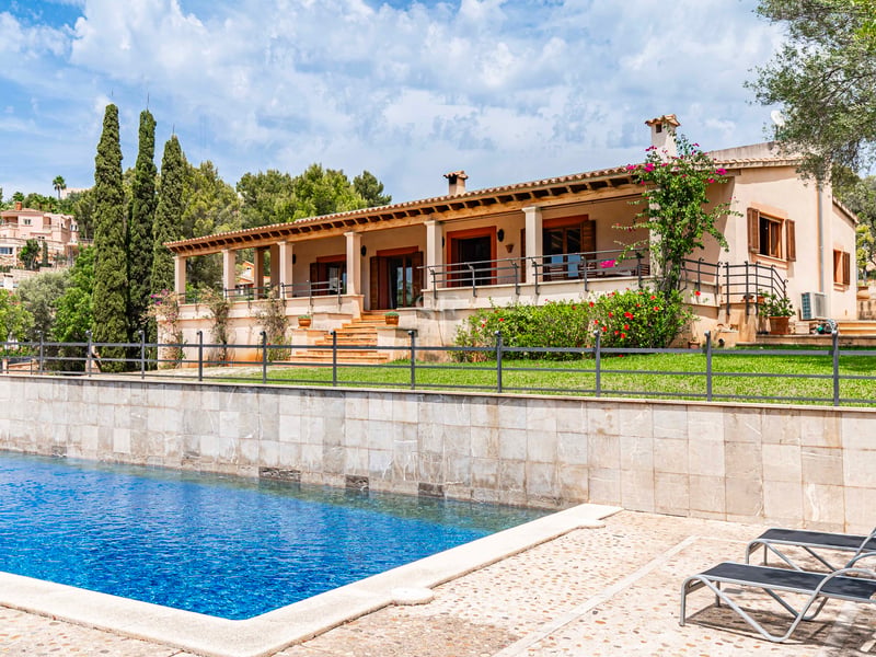 Grande piscine bleue et transats devant une villa méditerranéenne avec mur en pierre, pelouse et terrasse couverte.