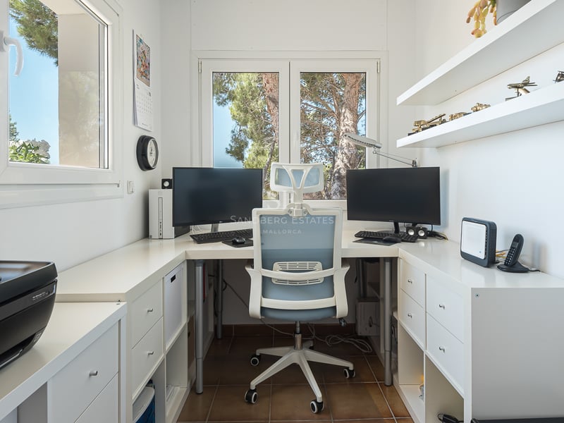 Bureau blanc moderne avec deux écrans, chaise ergonomique et fenêtres donnant sur des arbres verts.