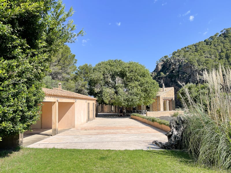 Large terrasse en pierre d'une villa méditerranéenne entourée d'arbres et de montagnes rocheuses sous un ciel bleu.