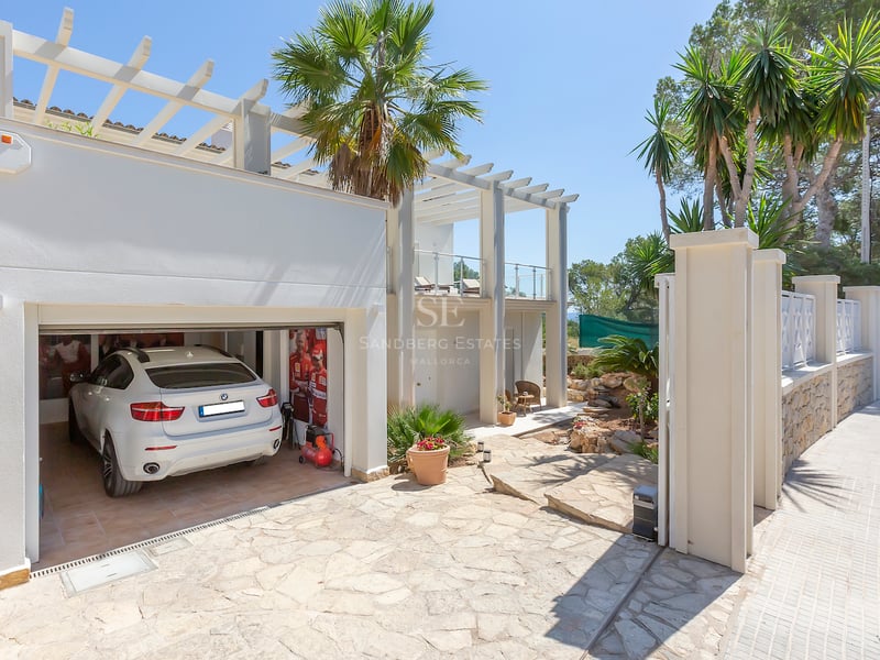 Extérieur d'une villa blanche avec allée en pierre naturelle, pergola et garage ouvert abritant une voiture de luxe.