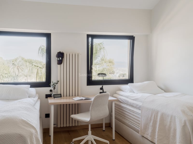 Chambre avec deux lits simples blancs, un bureau central et deux fenêtres donnant sur des palmiers.