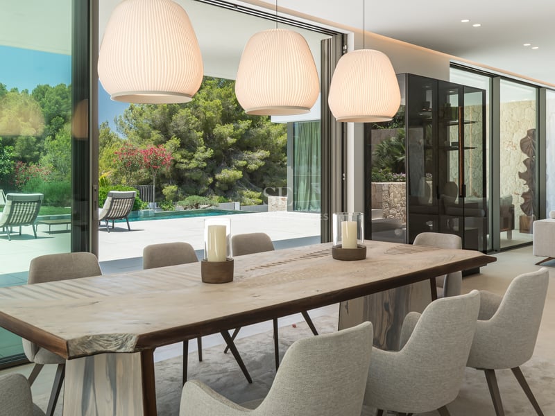 Salle à manger moderne avec table en bois naturel et chaises rembourrées donnant sur une piscine à travers des baies vitrées.