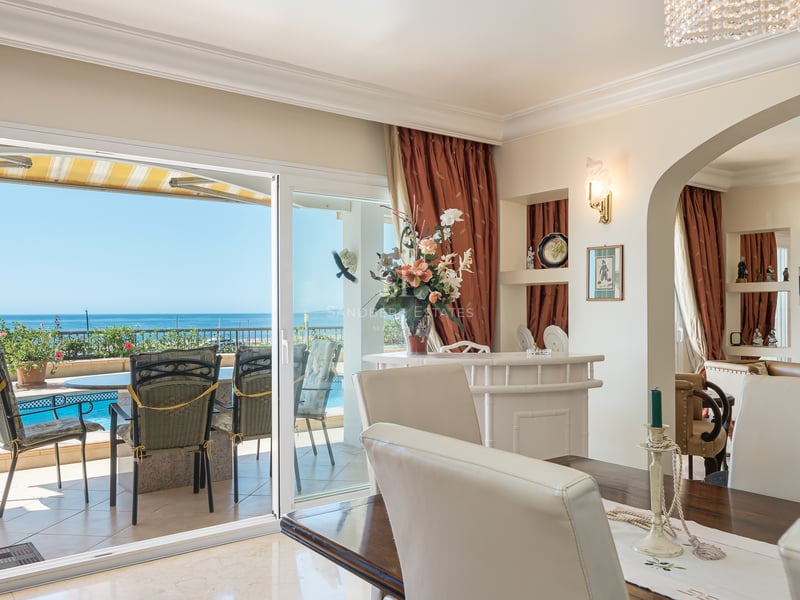 Salle à manger intérieure avec table en bois et chaises blanches donnant sur une terrasse avec piscine et vue mer.
