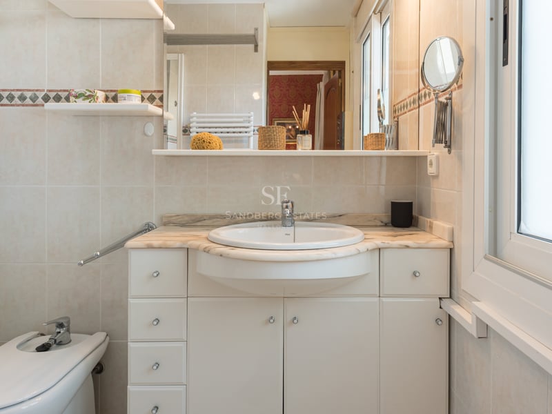 Vanité de salle de bain crème avec plan de travail en marbre, lavabo semi-encastré et bidet à côté d'une fenêtre.