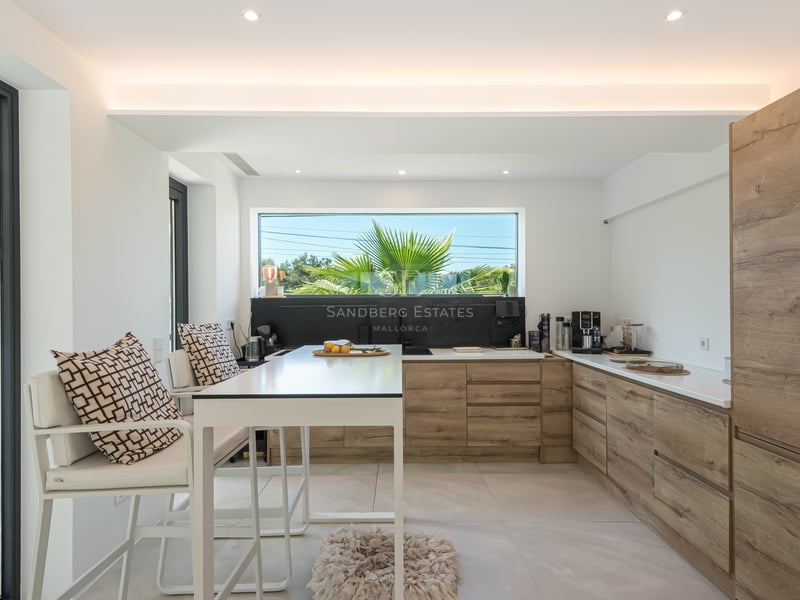 Cuisine contemporaine lumineuse avec des meubles en bois clair, un îlot blanc et une fenêtre cadrant des palmiers.