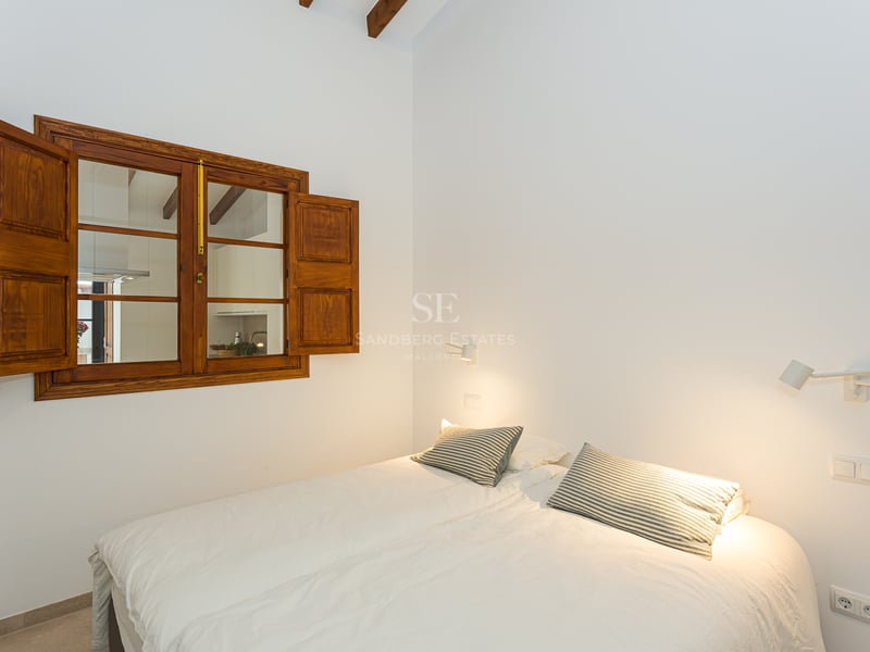 Chambre lumineuse avec literie blanche, oreillers rayés, fenêtre en bois avec volets et poutres apparentes.