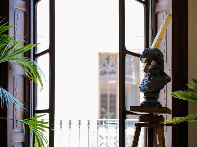 Escultura de un busto sobre un trípode de madera junto a un gran ventanal iluminado con plantas verdes.