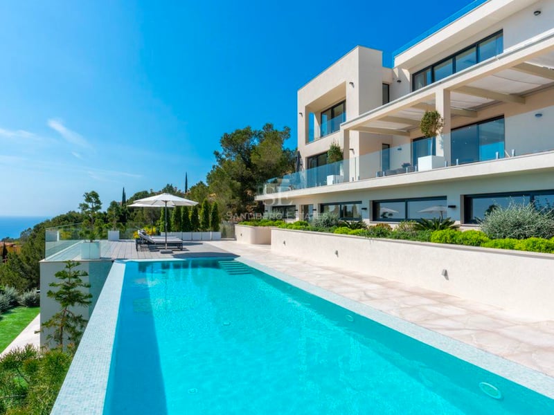 Villa moderne blanche avec piscine à débordement et vue sur mer sous un ciel bleu.