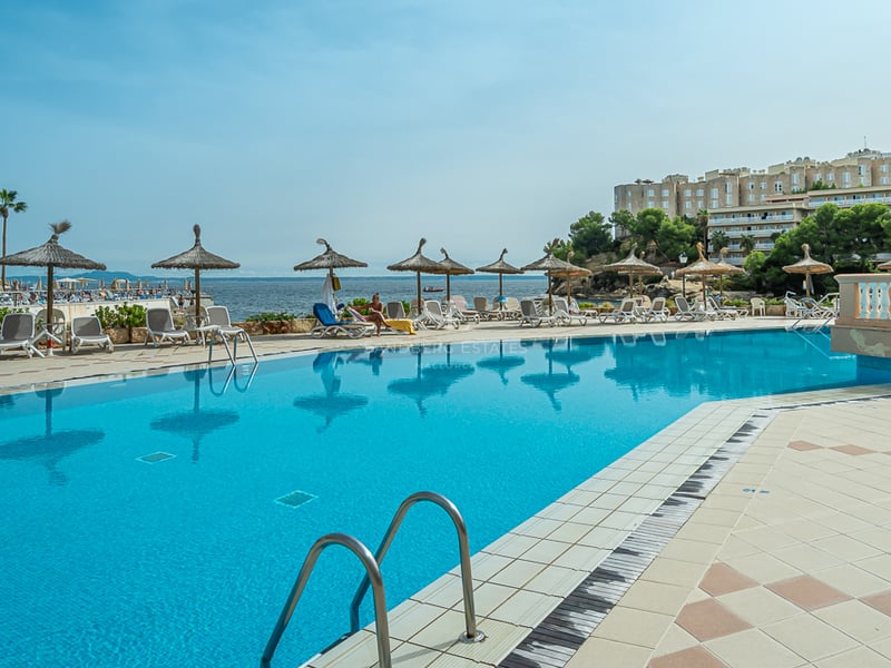 Grande piscine bleue entourée de transats et de parasols en paille avec vue sur la mer et les bâtiments côtiers.