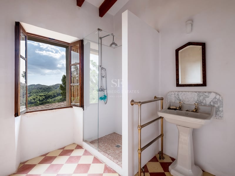 Salle de bain avec sol en damier, lavabo sur pied et douche vitrée près d'une fenêtre donnant sur les montagnes.