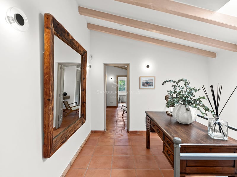 Couloir avec sol en terre cuite, grand miroir en bois, bureau antique et poutres apparentes au plafond.