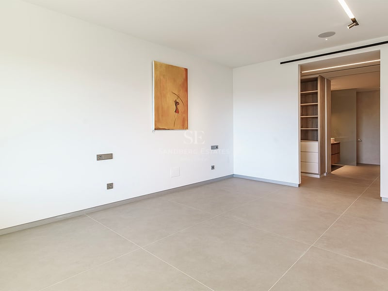 Chambre moderne vide avec grands carreaux beiges, murs blancs, art abstrait et vue sur un dressing en bois.