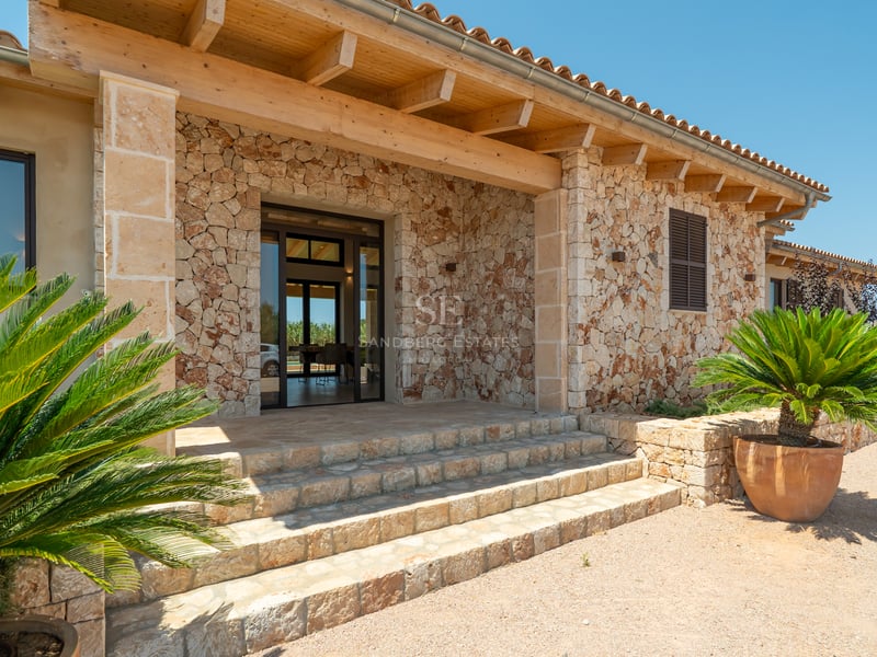 Fachada de piedra natural con vigas de madera, escalones de piedra y plantas mediterráneas bajo un cielo despejado.