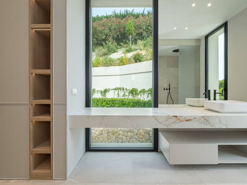 Salle de bains moderne avec plan de travail en marbre, vasque à poser, robinetterie noire et vue sur la végétation.