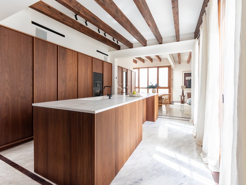Cocina moderna con isla de madera estriada, encimeras de mármol, vigas de madera vista y luz natural.