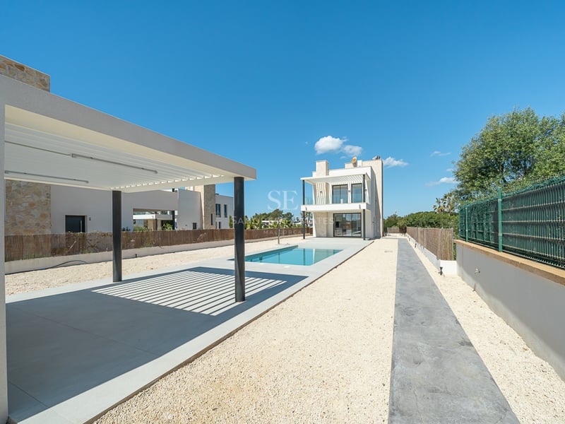 Extérieur d'une villa moderne avec piscine rectangulaire bleue, pergola blanche et cour en gravier sous un ciel clair.