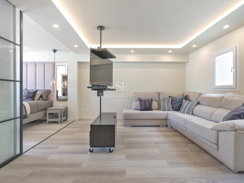 Salon contemporain avec grand canapé en L beige, support TV pivotant et chambre adjacente séparée par une paroi vitrée.