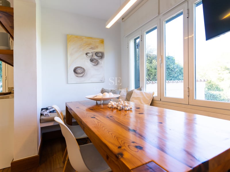 Coin repas avec une grande table en bois, chaises blanches et banquette avec coussins près d'une fenêtre ensoleillée.
