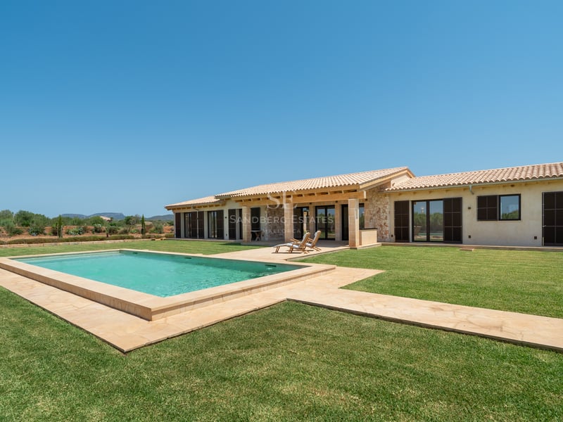 Piscina rectangular rodeada de césped frente a una villa de piedra de una planta bajo un cielo azul despejado.