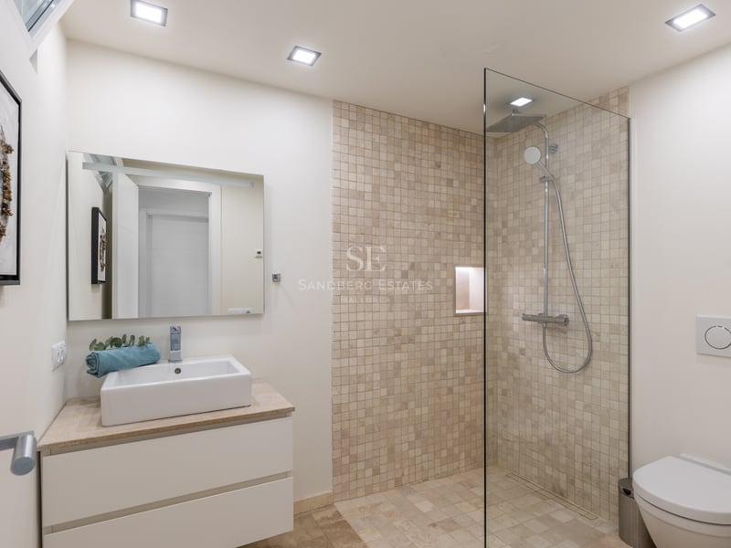 Salle de bains avec douche vitrée, mosaïque en pierre naturelle, meuble vasque blanc et finitions chromées.