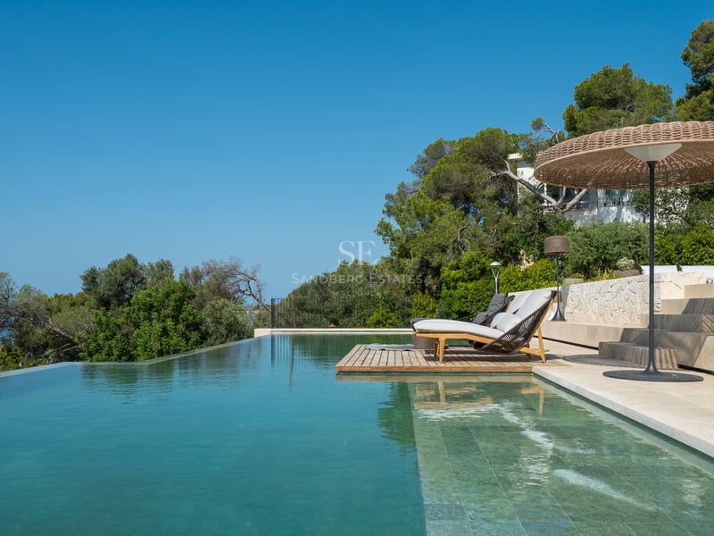 Piscine à débordement avec terrasse en pierre calcaire, bains de soleil et parasol tressé parmi les pins.