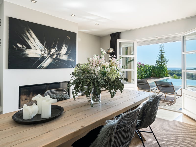 Salle à manger avec table en bois et cheminée ouvrant sur une terrasse avec piscine à débordement et vue mer.