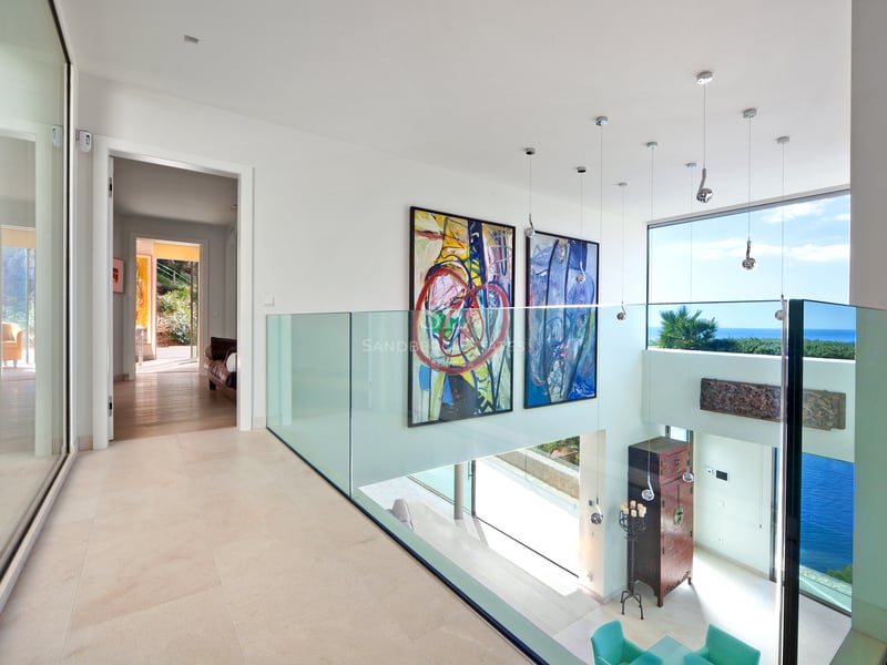 Couloir moderne avec garde-corps en verre, grands tableaux et vue sur la mer à travers de larges fenêtres.