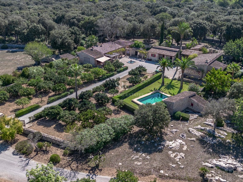 Vue plongeante d'un domaine en pierre avec piscine, palmiers et jardins luxuriants entourés de forêt.