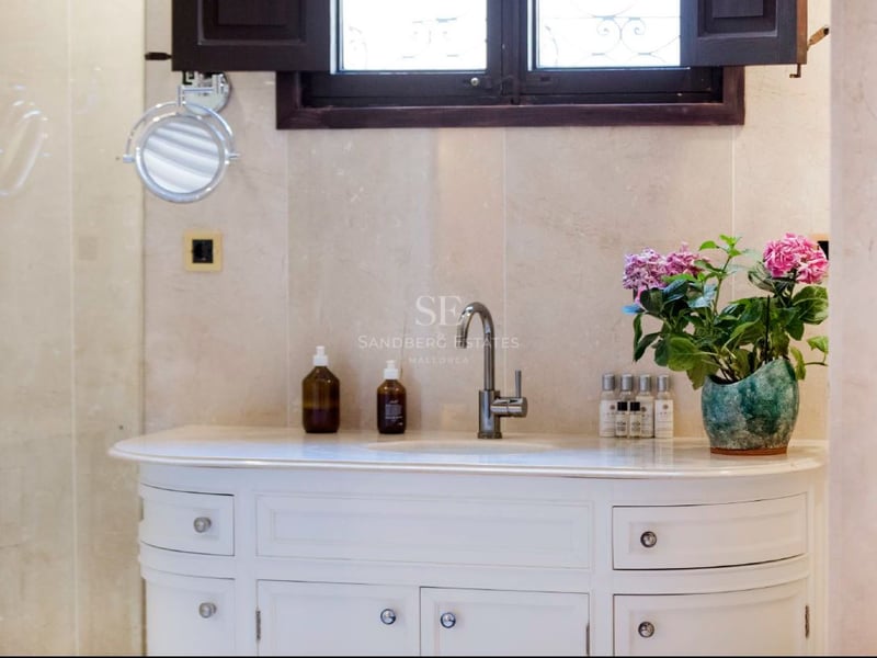 Lavabo blanco con grifo cromado, paredes de mármol beige, espejo de aumento y flores rosas.