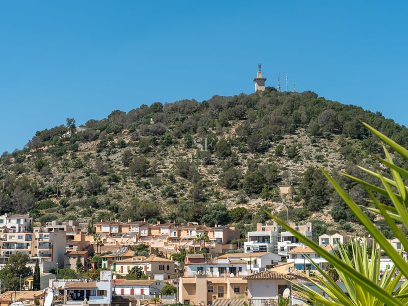 Vue de bâtiments résidentiels méditerranéens devant une colline verdoyante avec un monument sous un ciel bleu clair.