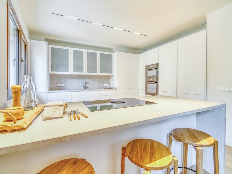 Cuisine minimaliste lumineuse avec placards blancs, îlot en pierre avec tabourets en bois et appareils intégrés.